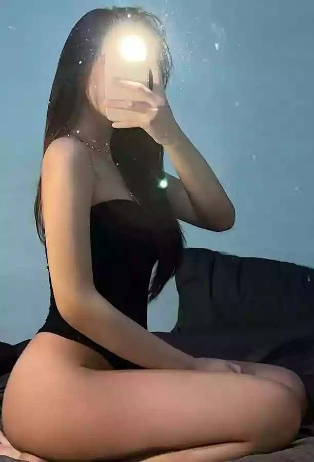 Person wearing a black one-piece swimsuit sits cross-legged on a bed, taking a mirror selfie with a phone obscuring their face.