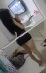 Person standing in a room taking a mirror selfie with a smartphone, wearing a black outfit; a second person is on the floor nearby in the background.