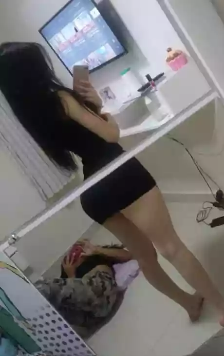 Person standing in a room taking a mirror selfie with a smartphone, wearing a black outfit; a second person is on the floor nearby in the background.