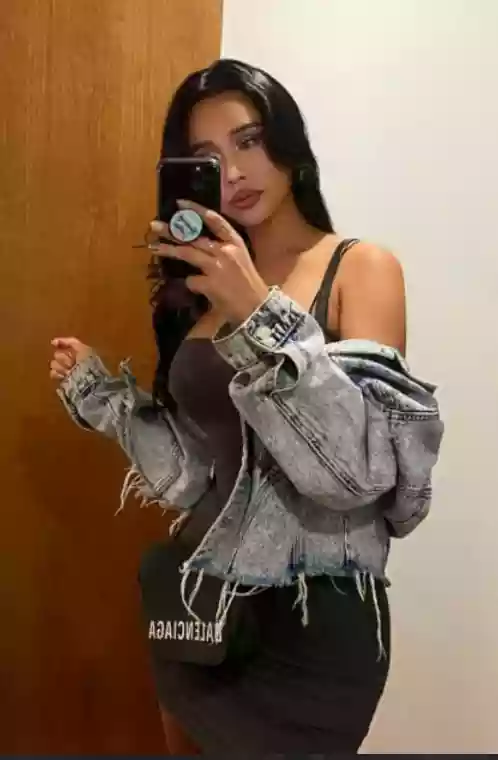 Woman posing for a mirror selfie in a doorway, wearing a distressed gray denim jacket over a dark off-shoulder dress and holding a smartphone with a round pop socket visible on the back