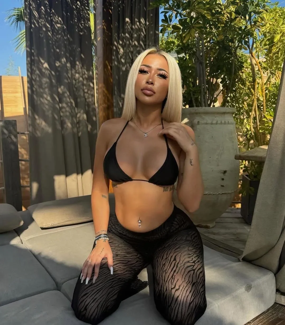 Woman posing outdoors on a gray couch in a black bikini top and patterned black leggings, sunlit backyard plants in the background.