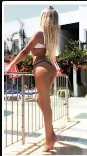 Woman in a white bikini standing at a poolside railing, back to camera in a sunlit courtyard.