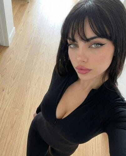 Self-portrait of a woman with dark hair and winged eyeliner wearing a black V-neck top, standing in a light wood-floored hallway and looking up at the camera.