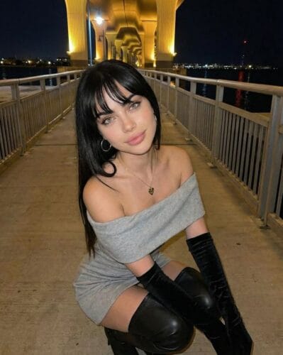 Young woman kneeling on a night-lit bridge with yellow arches in the background, wearing a gray off‑the‑shoulder dress and long black gloves.