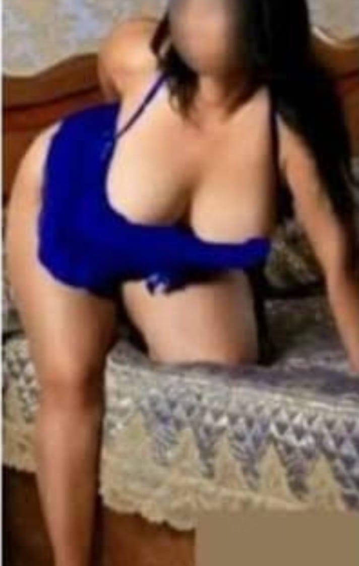 Woman in a bright blue top leaning on a bed, showing cleavage.